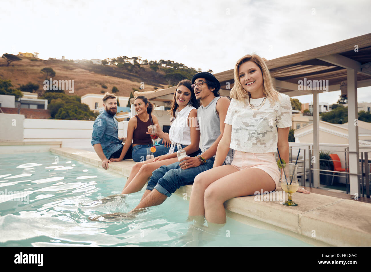 Lächelnde junge Frau Blick in die Kamera mit ihren Freunden sitzen am Rand des Swimming Pool. Junge Menschen genießen auf der Dachterrasse Partei Stockfoto