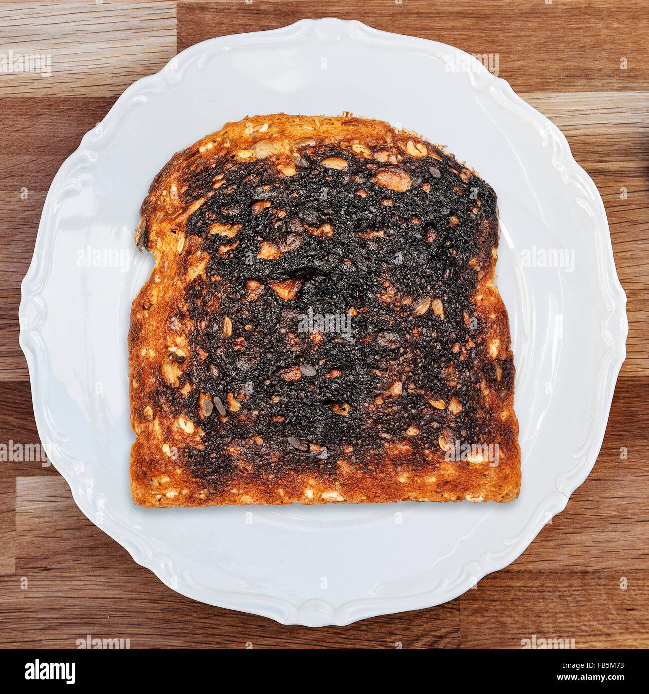 Ein Stück von verbranntem Toast auf einem glänzend weißen Teller auf einer hölzernen Tischplatte gelegt. Stockfoto