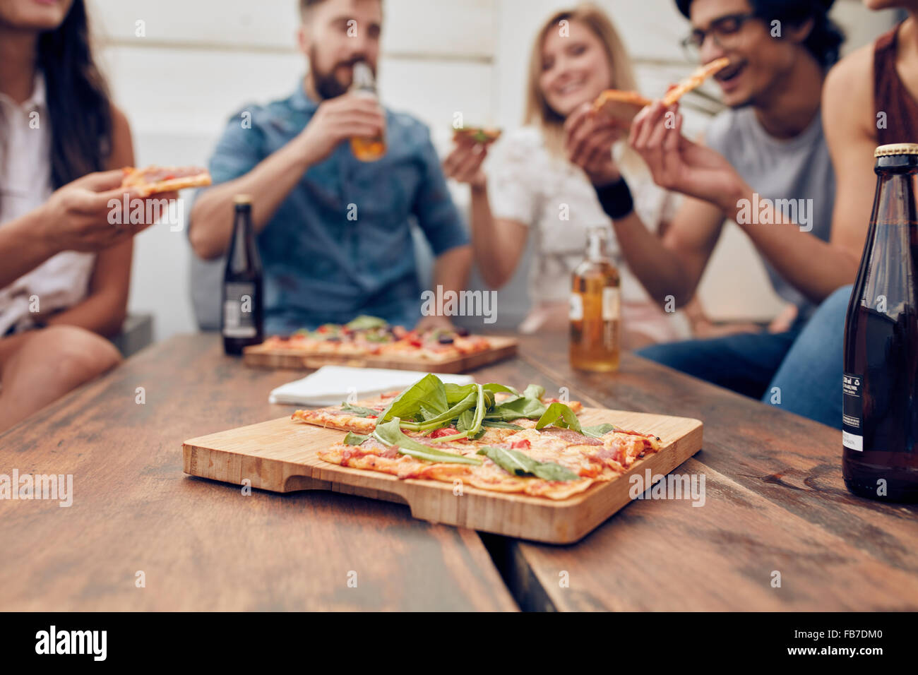 Schuss von Pizza auf Holzplatte mit Menschen Essen und trinken im Hintergrund hautnah. Gruppe von Freunden gesammelt, um den Tisch herum Stockfoto