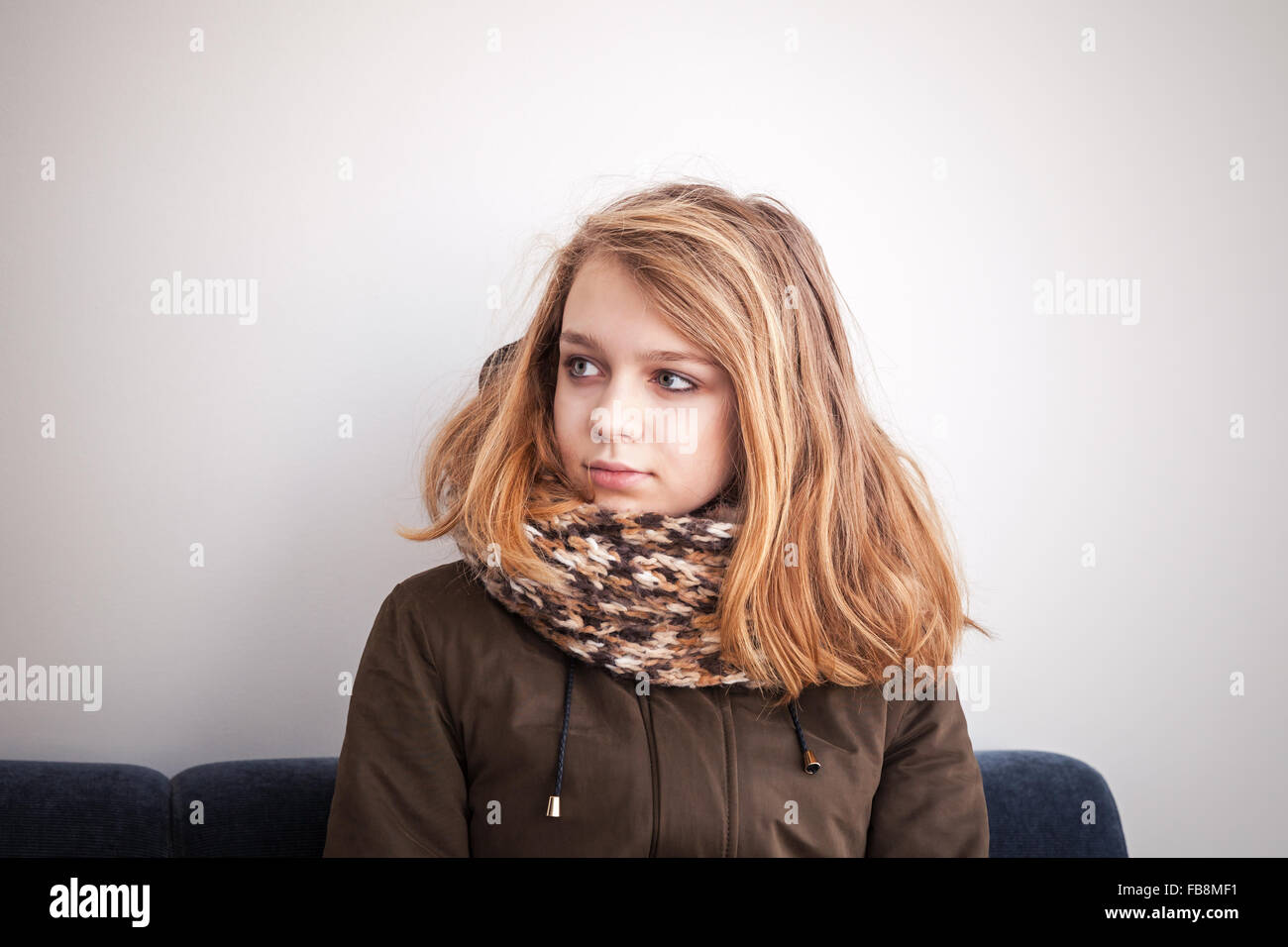 Schöne blonde kaukasische Teenager-Mädchen in warmen outwear sitzen auf blauem Sofa über weiße Wand Stockfoto