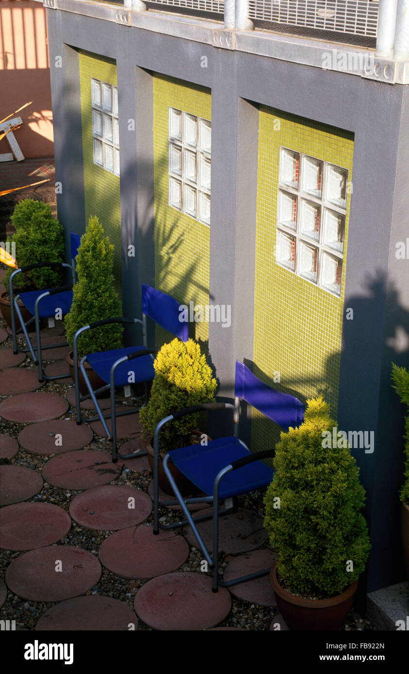 Terrakotta geflieste Terrasse mit blauem Segeltuch Stühle und Töpfe von Nadelbäumen gegen gelb gefliesten Wand mit Glasfenstern Ziegel Stockfoto