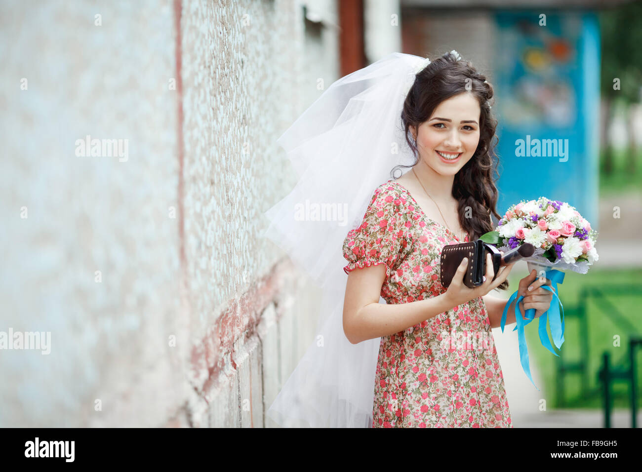 Die Braut in einem schlichten Retro-Kleid mit Blumenmuster, bereits tragen Schleier, Brautstrauß und Handtasche, posiert vor Haus, direkt in die Kamera, lächelnd. Stockfoto