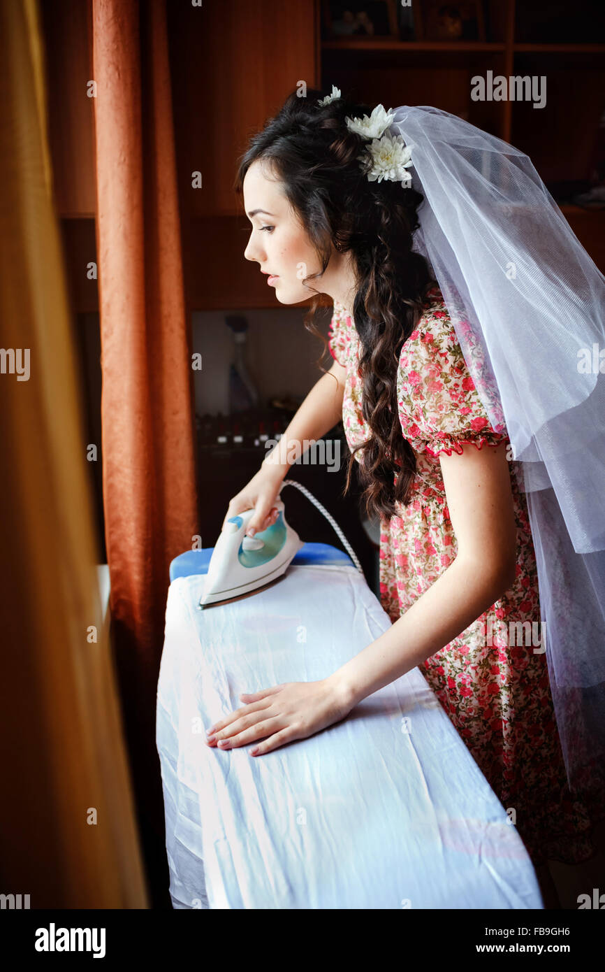 Die Braut, Vorbereitung für Trauung, Bügeln ihr Kleid an Bord, natürliches Licht. Sie trägt ein buntes retro Kleid Blick Fenster an Zukunft denken. Stockfoto