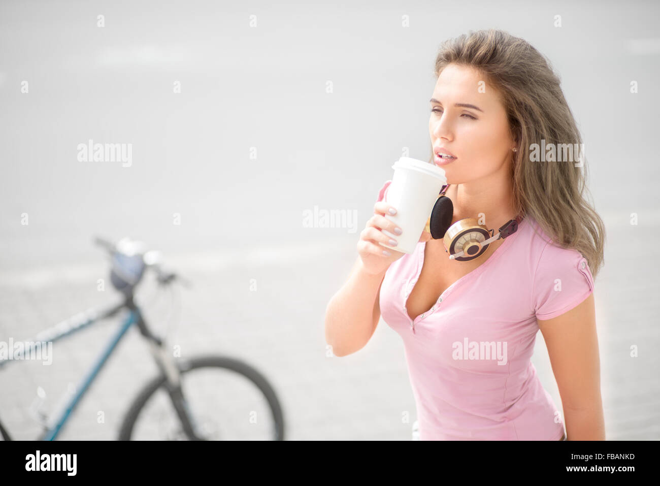 Schöne junge Frau trägt Musik Kopfhörer um den Hals, trinken Kaffee aus einem Take away Kaffee Becher gegen Fahrrad und Stockfoto