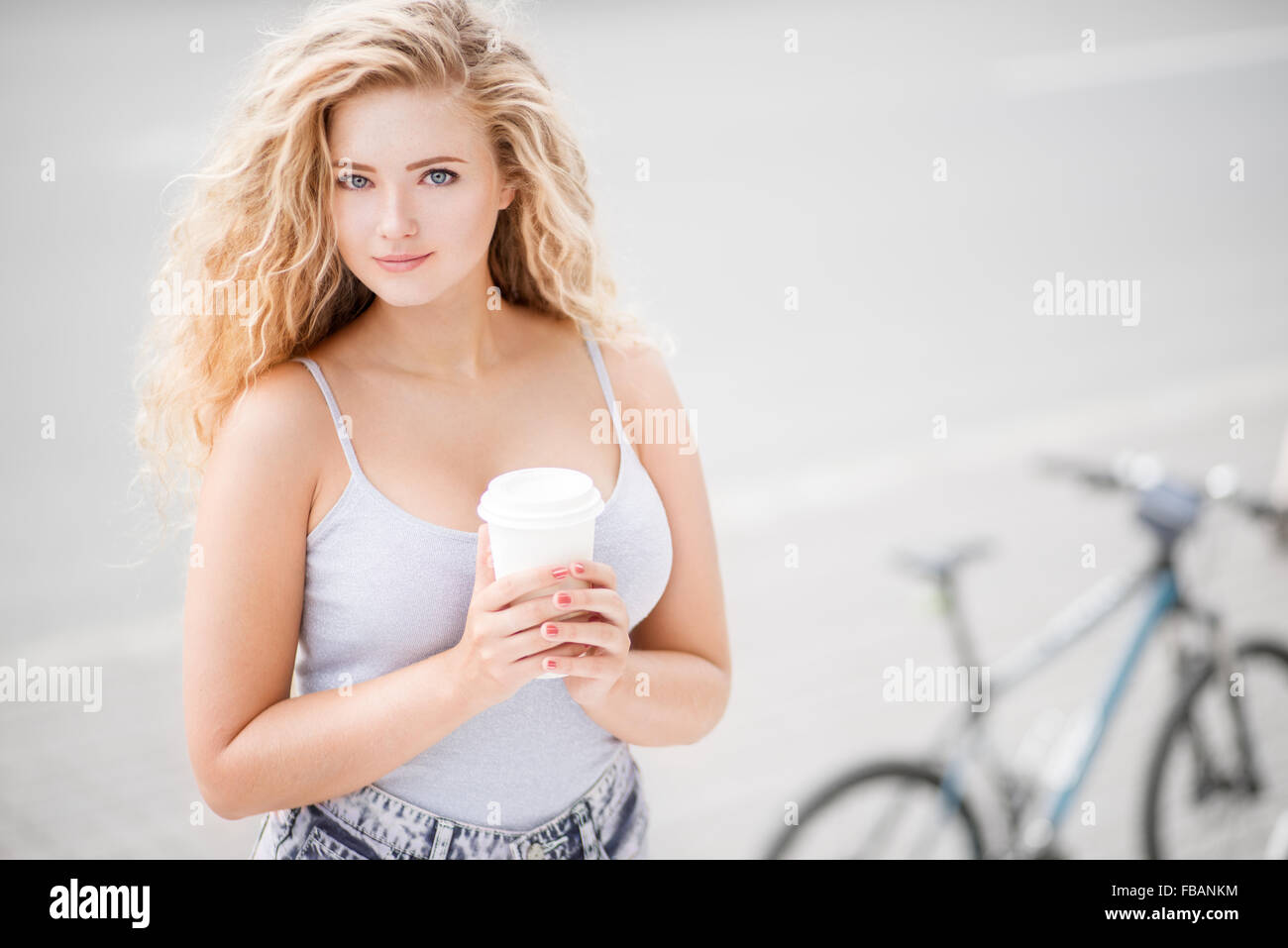 Glückliche junge Frau mit langen lockigen Haaren, ein Take away Kaffeetasse halten und gegen leere Straße und Stadt-Fahrrad-Hintergrund. Stockfoto