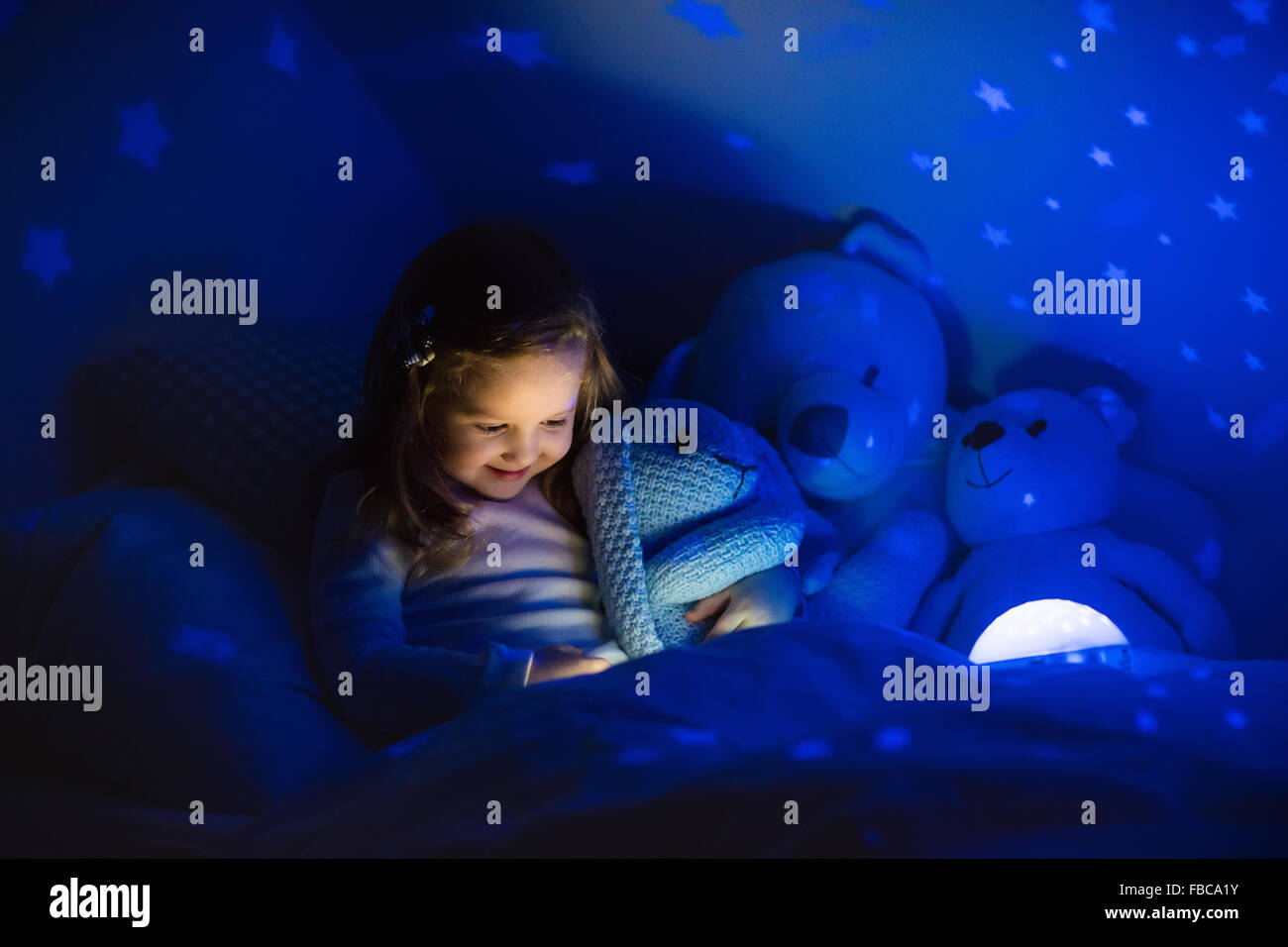Kleines Mädchen ein Buch im Bett lesen. Dunklen Schlafzimmer mit Nachtlicht Sterne auf Decke zu projizieren. Kinder Kindergarten und Bettwäsche. Stockfoto