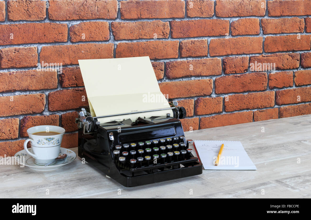 Vintage China, Schreibmaschine, Notizblock und Bleistift Teetasse mit Tee auf alten schäbigen Holzeffekt Tischplatte mit rotem Backstein-Mauer-Hintergrund Stockfoto