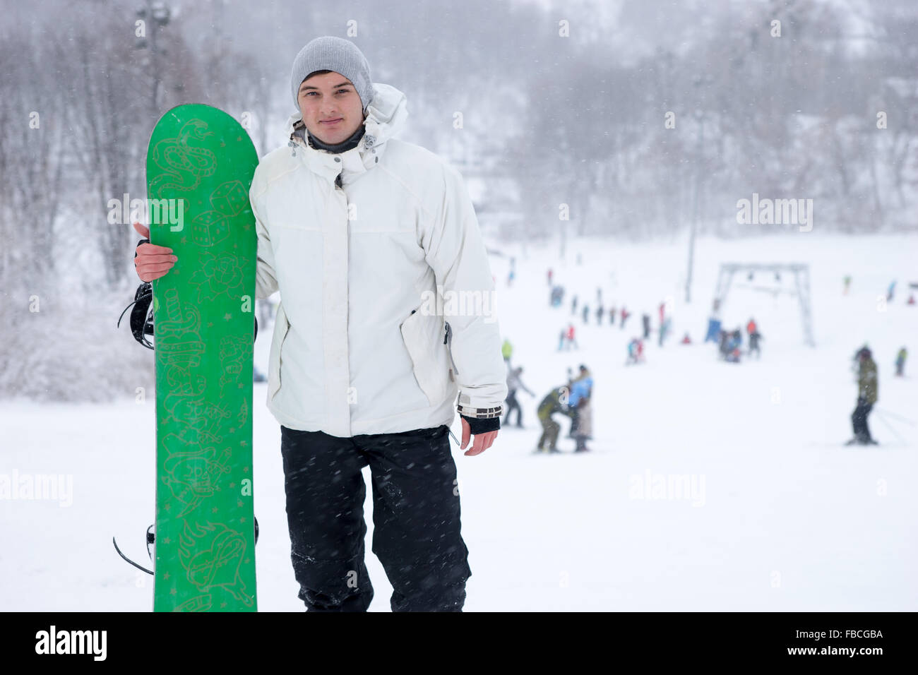 Sportlicher junger Mann hält ein helles Grün Snowboard auf einem verschneiten Berg laufen in einem Skigebiet posiert für die Kamera mit einem Lächeln Stockfoto