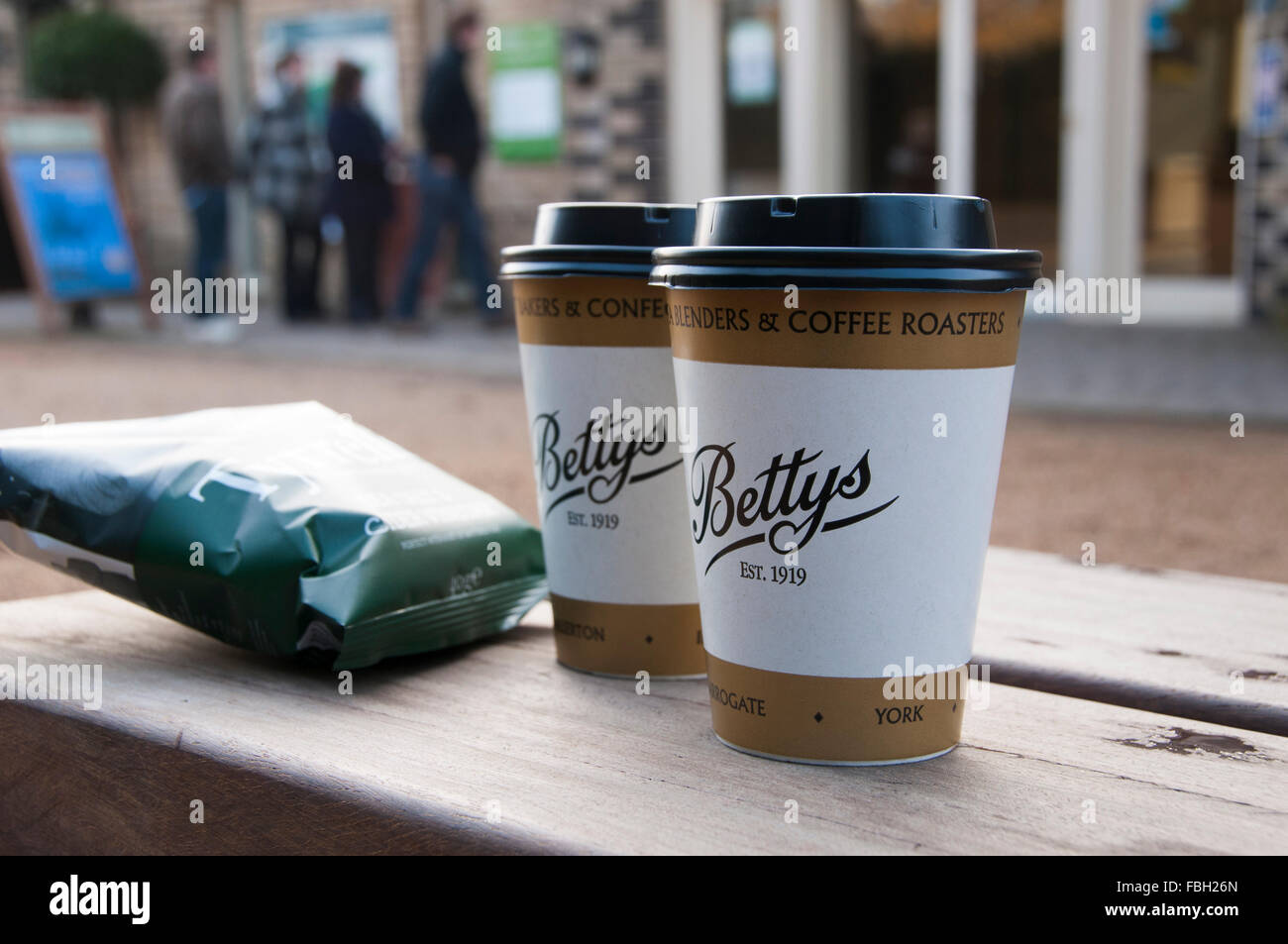 2 zum Mitnehmen Tassen (Deckel auf) und ein knackig-Paket, auf einem hölzernen Tisch außen Bettys Cafe an der RHS Harlow Carr, Harrogate, North Yorkshire, England, GB, UK. Stockfoto