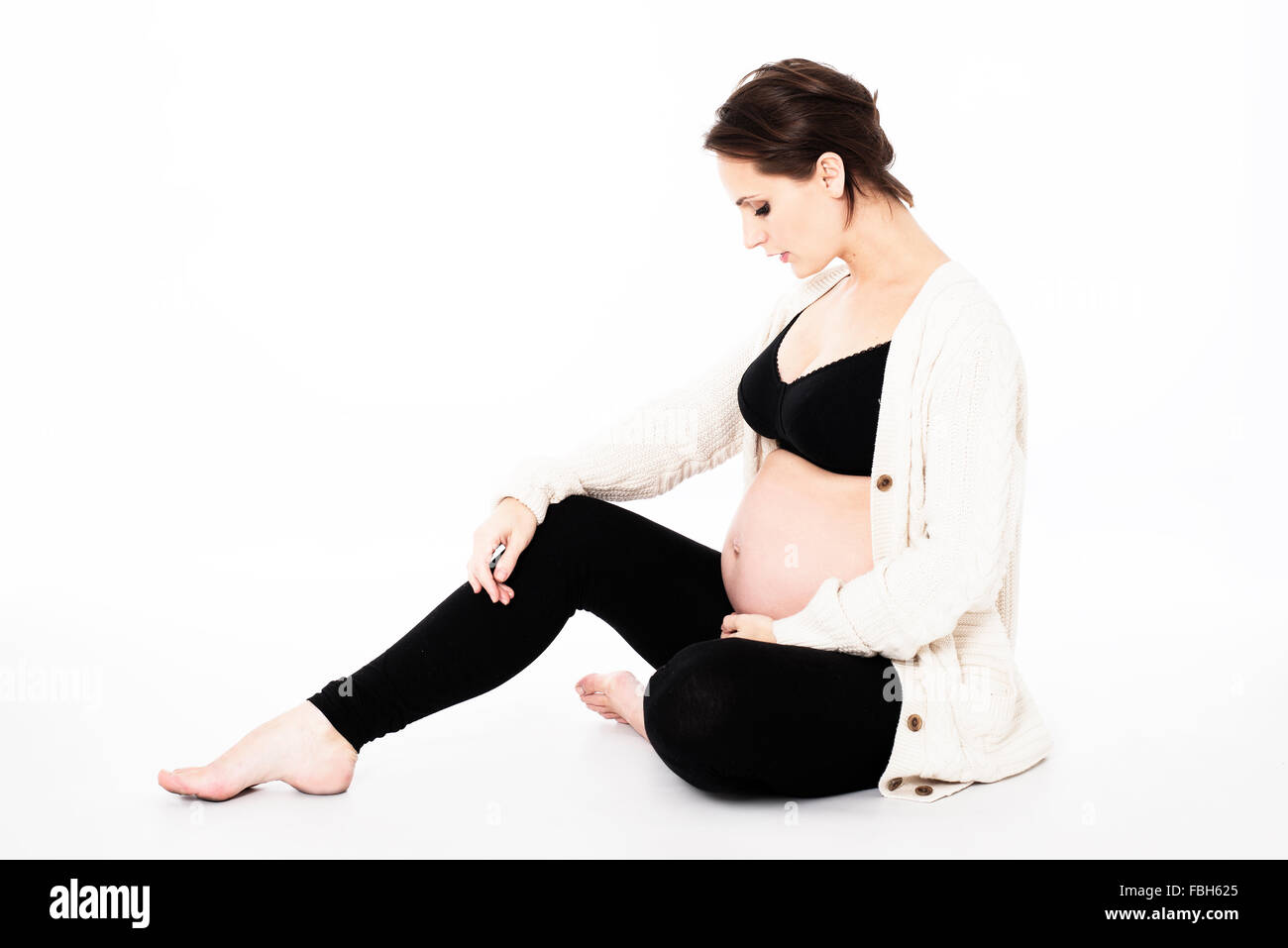 schöne schwangere Frau in schwarzen Leggins und Sport-BH mit weißer Wolle Strickjacke auf Boden sitzend, stoßen auf Show Stockfoto