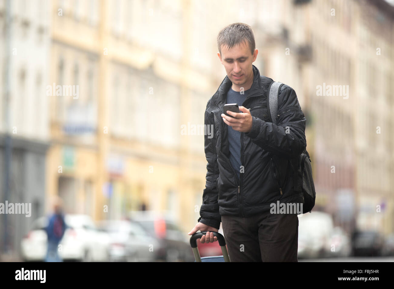 Junger attraktiven Mann mit Gepäck lächelnd Tasche auf der regnerische Stadtstraße mit Blick auf Smartphone, app, Gps, mit Suche Stockfoto