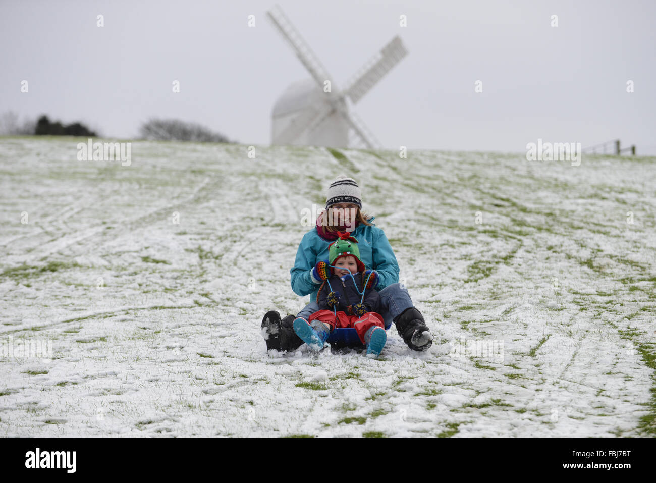 Eine Mutter und seinem Sohn auf einem Schlitten sitzen zusammen und schieben Sie eine verschneite Hügel vor einem der Clayton Windmühlen in Clayton, East Sussex, UK. Stockfoto