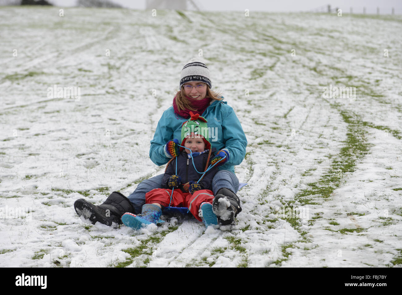Eine Mutter und seinem Sohn auf einem Schlitten sitzen zusammen und schieben Sie eine verschneite Hügel vor einem der Clayton Windmühlen in Clayton, East Sussex, UK. Stockfoto