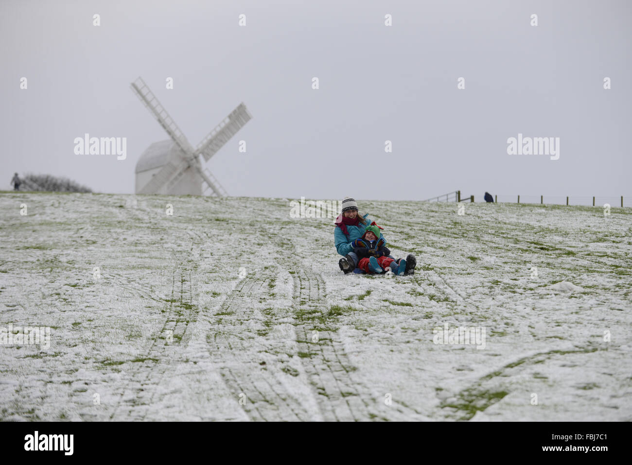 Ein Vater und sein Sohn auf einem Schlitten sitzen zusammen und schieben Sie eine verschneite Hügel vor einem der Clayton Windmühlen in Clayton, East Sussex, UK. Stockfoto