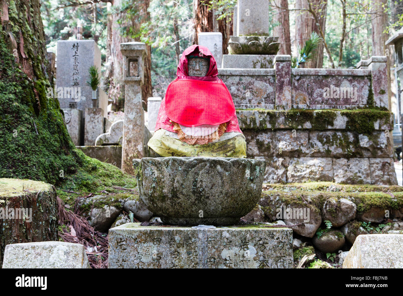 Japan, Koyasan, berühmte Okunoin Friedhof. Japanische Jizo Statue mit roter Latzhose und Hut und grünen Rock, sitzen auf den Stein Lotus flower Sockel auf dem Grab. Stockfoto