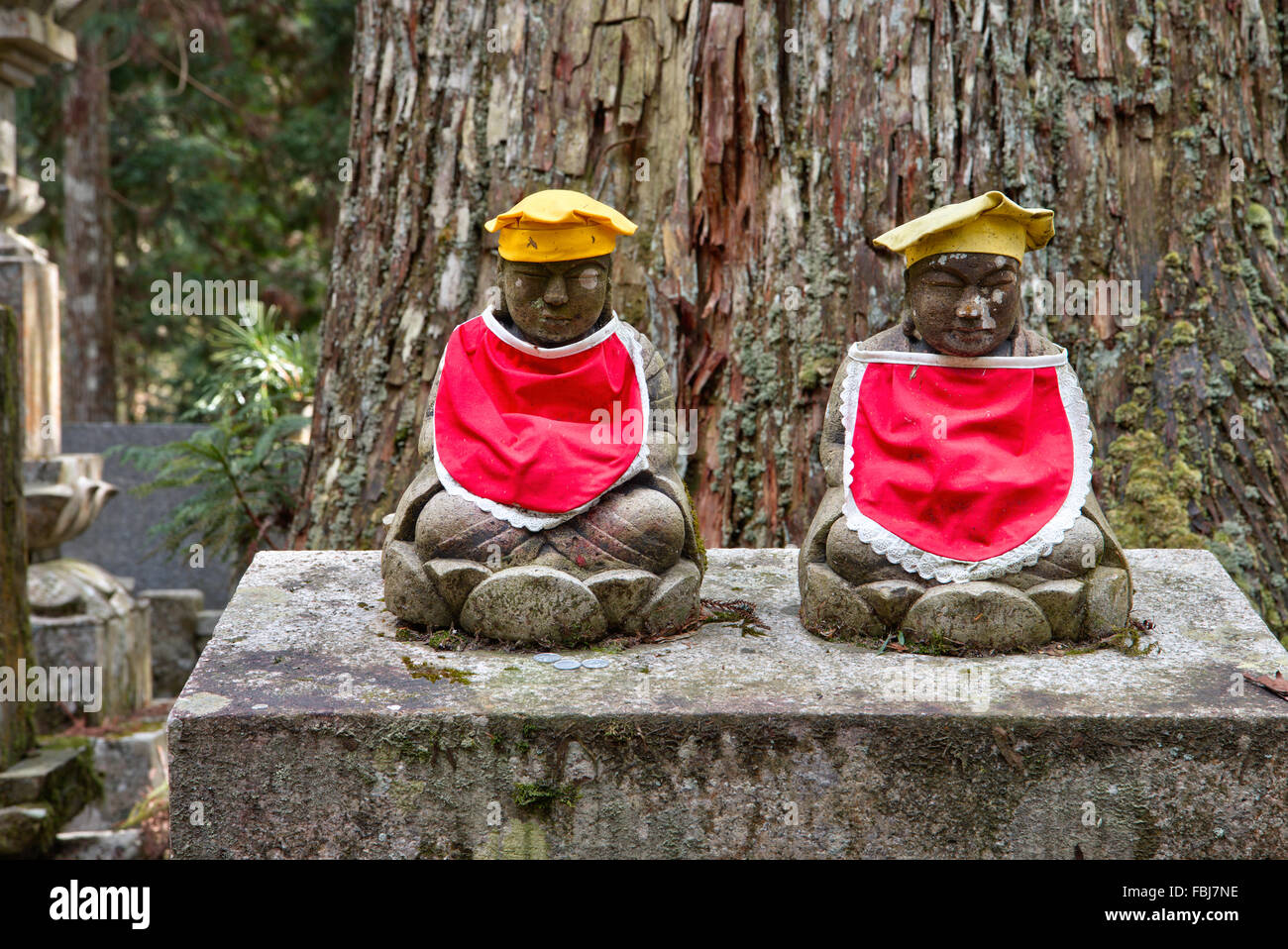 Japan, Koyasan, berühmte Okunoin Friedhof. Zwei kleine japanische Jizo Statuen mit red Bibs und gelbe Hüte sitzen nebeneinander auf Grabstein. Stockfoto