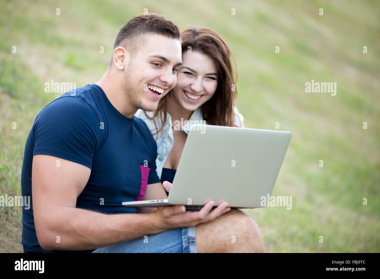 Porträt von positiven jungen Mann und schöne Frau tragen legere Kleidung auf Sommertag auf dem Rasen im Park sitzend, auf Schoß arbeiten Stockfoto