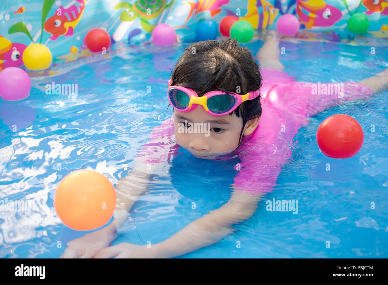Ein kleines Mädchen in rosa Anzug spielt Wasser und Kugeln in blau Planschbecken Stockfoto