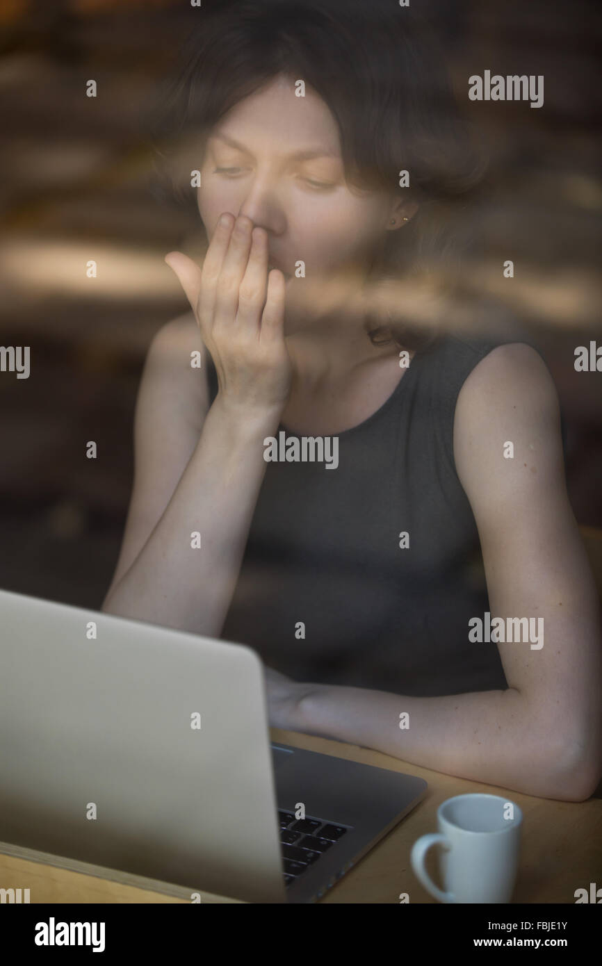 Junge Frau sitzt im Cafe gegenüber Laptop mit Tasse Kaffee, Gähnen, Blick auf Bildschirm, faul zu früh arbeiten Stockfoto