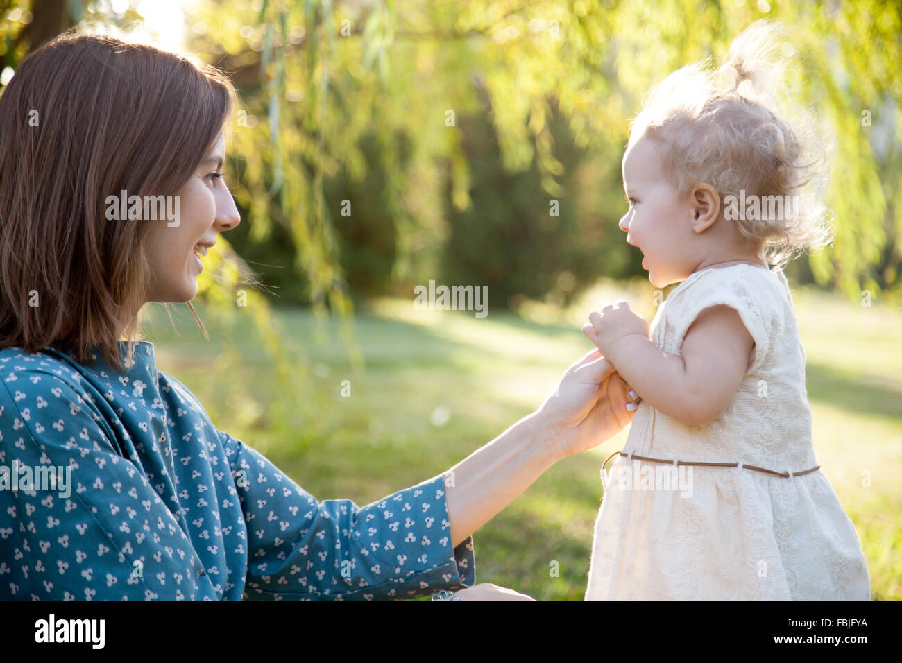 Glückliche junge Mutter und entzückende blonde Mädchen spielen zusammen im Park im Sommer, lächelnde Mutter ihre kleine Tochter Hand haltend, Stockfoto