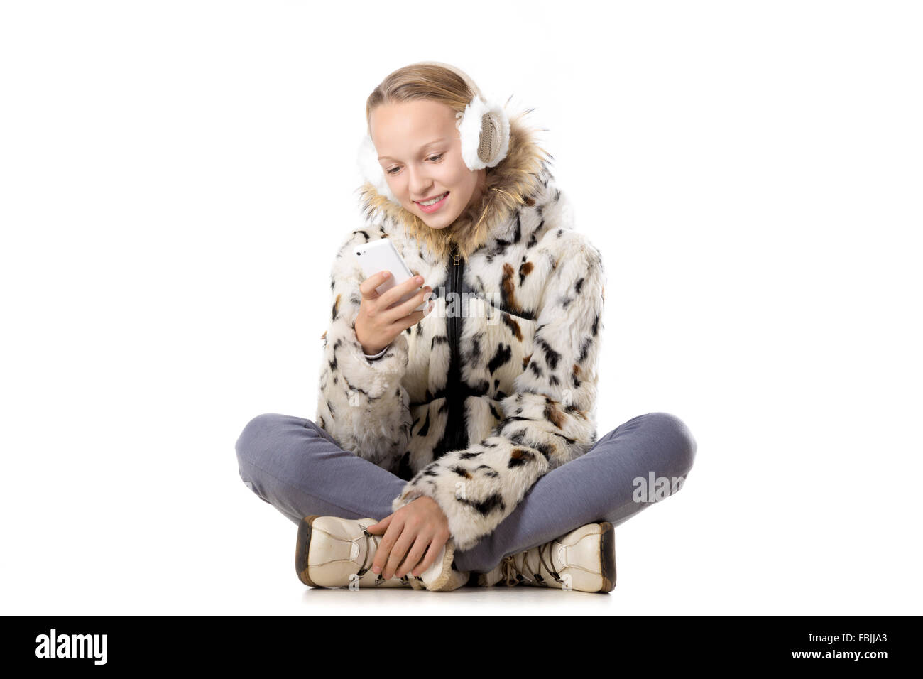 Glücklich lächelnd schön lässig Teenager-Mädchen tragen Wintermantel und Ohrenschützer, sitzt im Schneidersitz, Handy in den Händen halten Stockfoto