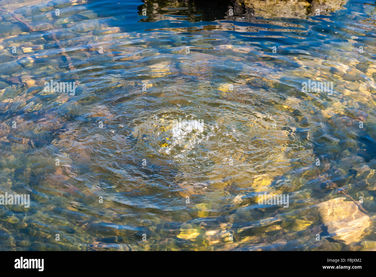 Klares Wasser sprudeln in kleinen outdoor Teich, bilden konzentrische Wellen, mit grünen Felsen und Steine unter Wasser sichtbar. Stockfoto