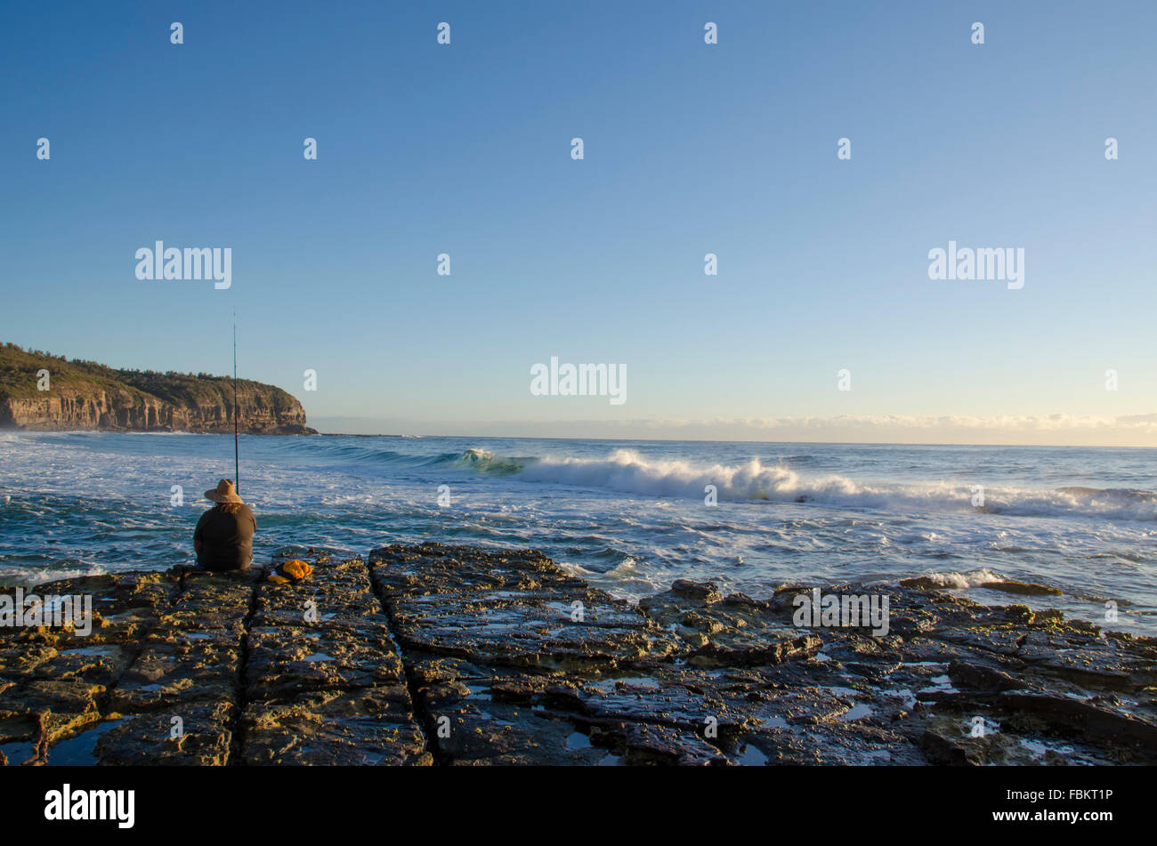 Eine kaukasische Person fischt in der Morgendämmerung am Rand einer felsigen Plattform, während Wellen hereinrollen und die Sonne an einem Strand von Sydney aufgeht Stockfoto
