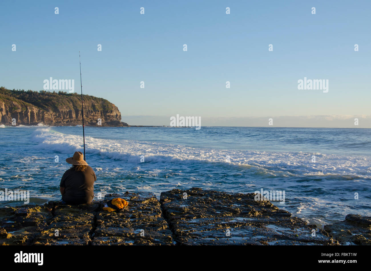 Eine kaukasische Person fischt in der Morgendämmerung am Rand einer felsigen Plattform, während Wellen hereinrollen und die Sonne an einem Strand von Sydney aufgeht Stockfoto