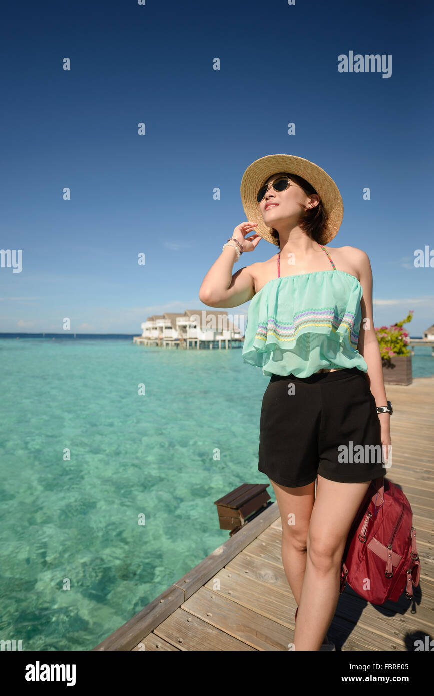 schlanke Frau mit Hut und Sonnenbrille posting auf Holzbrücke über dem kristallklaren Meerwasser auf den Malediven an einem sonnigen Tag Stockfoto