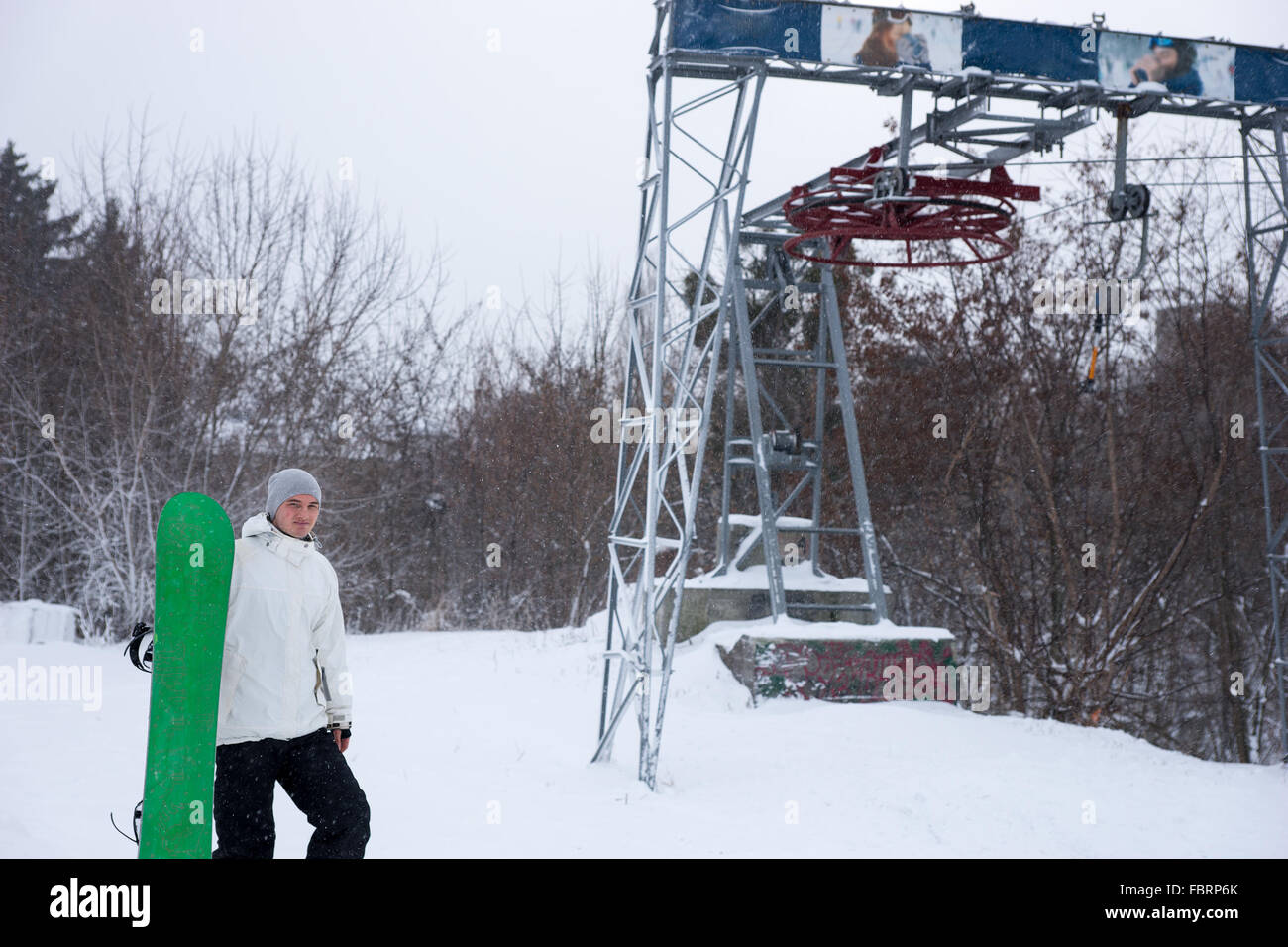 Einzelne sportlicher Mann im weißen Kittel trägt ein grünes Snowboard auf Ski Resort Hügel neben Hubwerk im winter Stockfoto