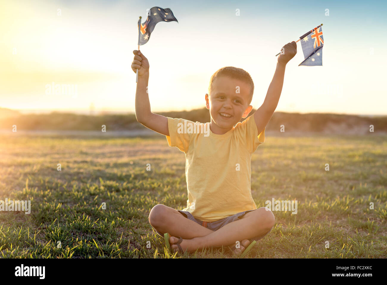 Fröhlicher Junge mit Flaggen Australiens sitzen auf dem Rasen bei Sonnenuntergang am Australia Day Stockfoto