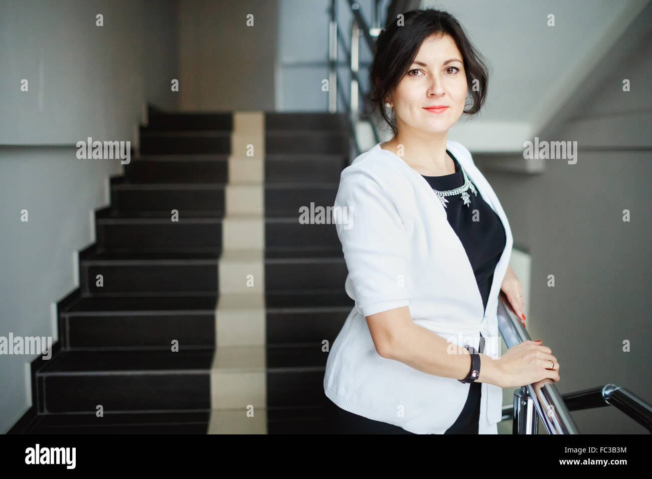 Erfolgreiche attraktive Brünette mit freundlichen Augen stehend auf der Treppe des Bürogebäudes, während einer Pause bei der Arbeit. Weiße Jacke und strengen schwarzen Kleid Schmuck tragen. Nette junge Frau posiert. Stockfoto