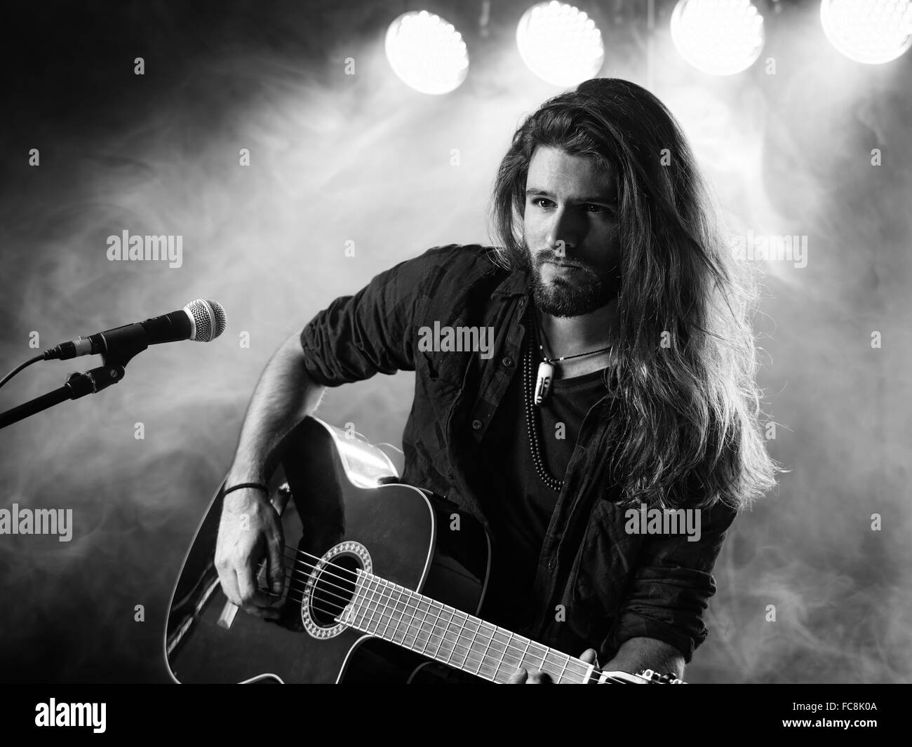 Foto von einem jungen Mann mit langen Haaren und Bart spielt eine akustische Gitarre auf der Bühne mit Lichtern und Konzertatmosphäre. Stockfoto