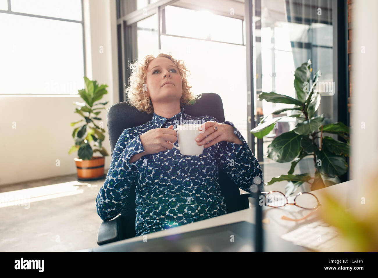 Geschäftsfrau sitzt an ihrem Schreibtisch im Büro mit einer Tasse Kaffee wegschauen und denken. Stockfoto