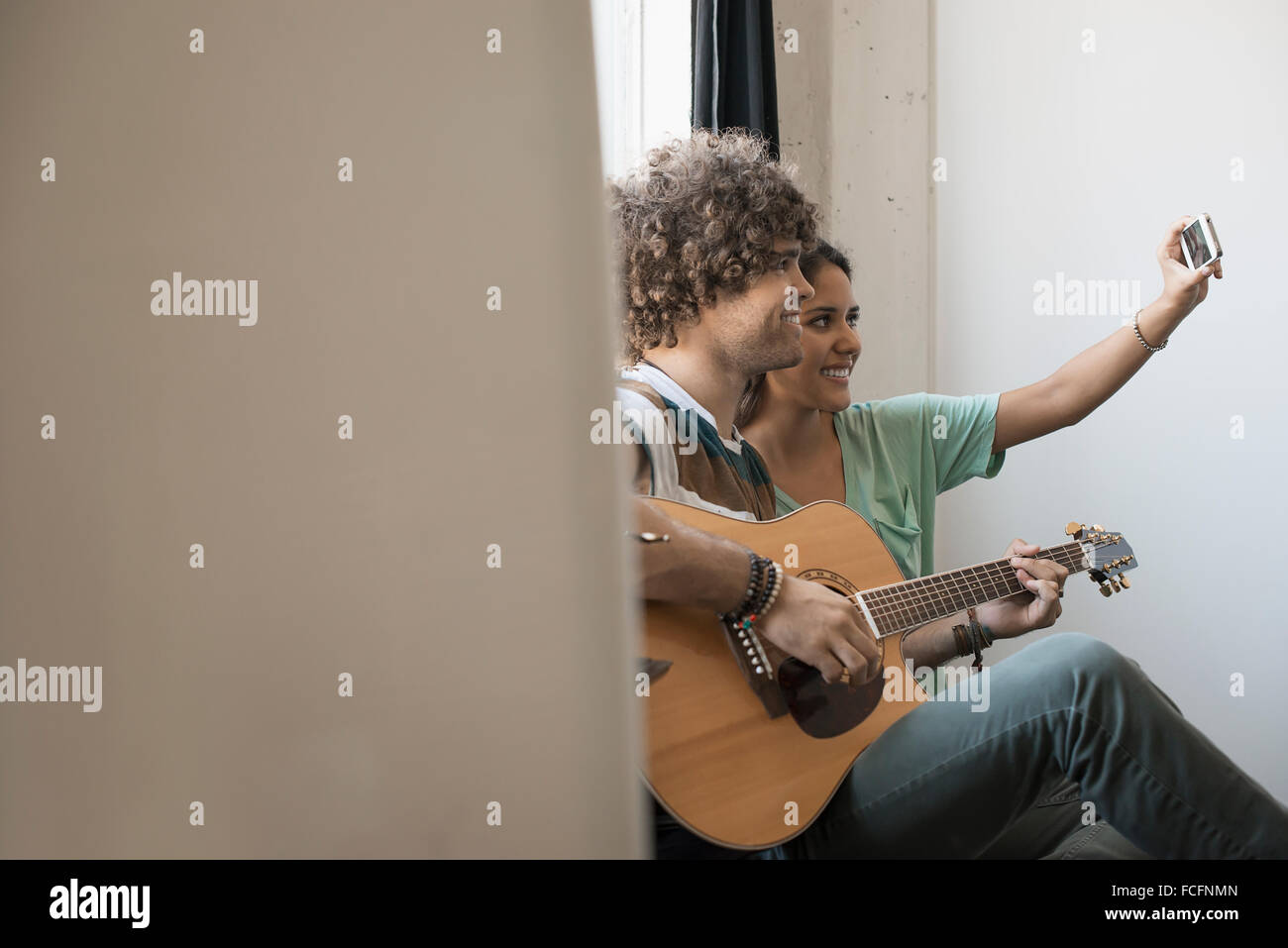 Loft-lebendigen. Ein junger Mann spielt Gitarre und eine Frau neben ihm unter einem selfy mit einem Smartphone. Stockfoto