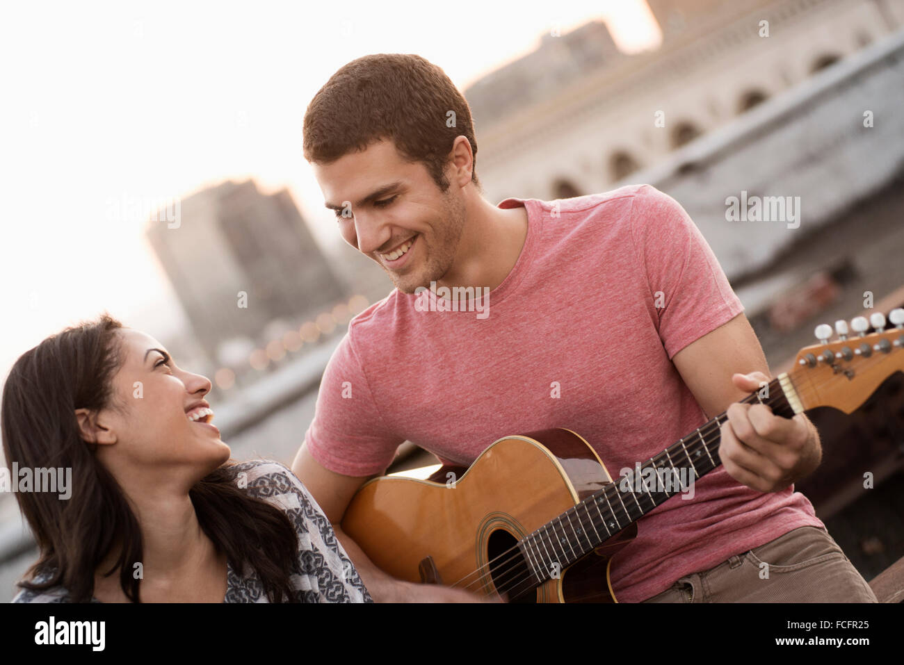 Ein Mann, die Gitarre zu einer Frau auf einer Dachterrasse mit Blick auf eine Stadt in der Dämmerung. Stockfoto