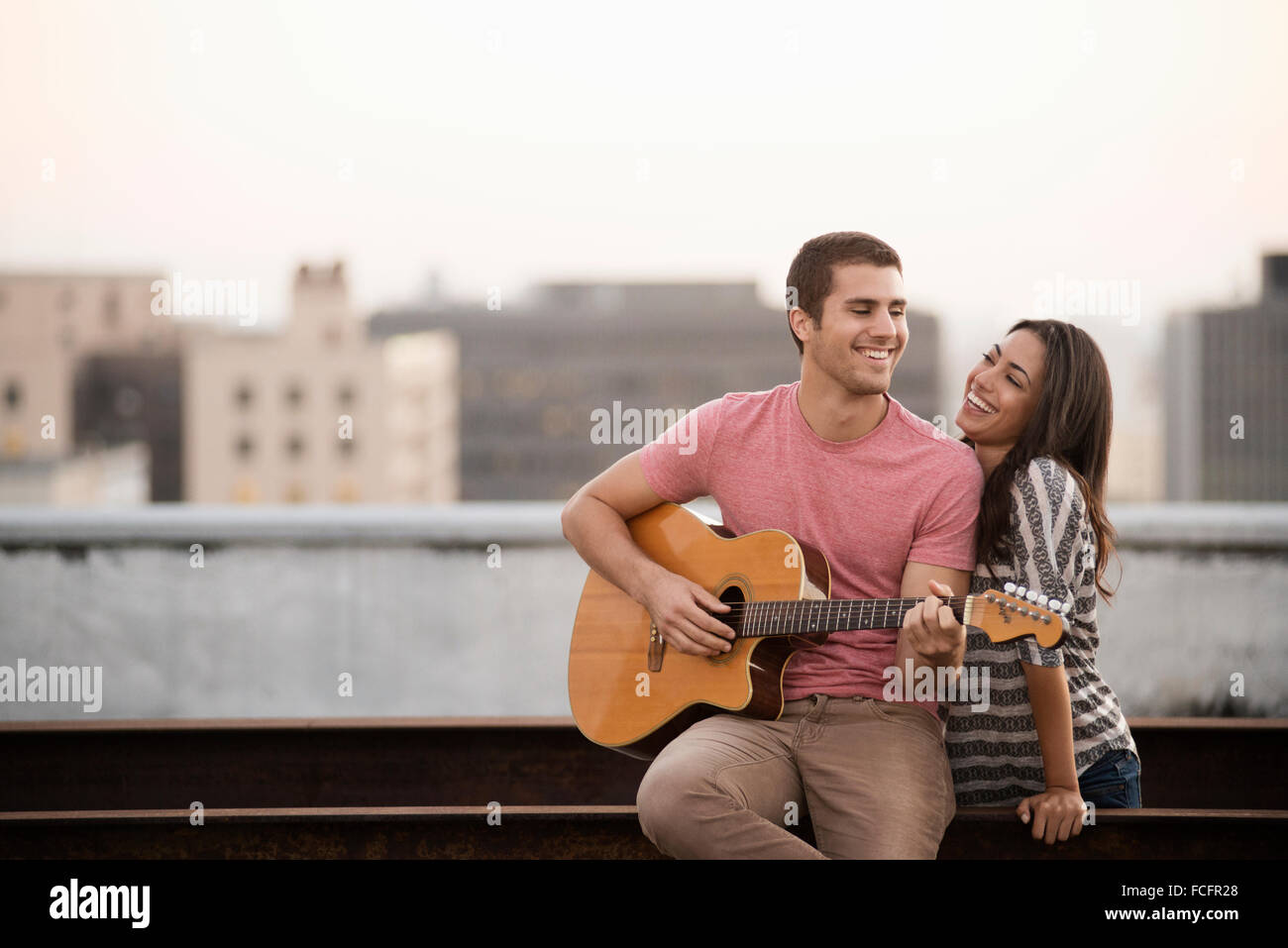 Ein Mann, die Gitarre zu einer Frau auf einer Dachterrasse mit Blick auf eine Stadt in der Dämmerung. Stockfoto