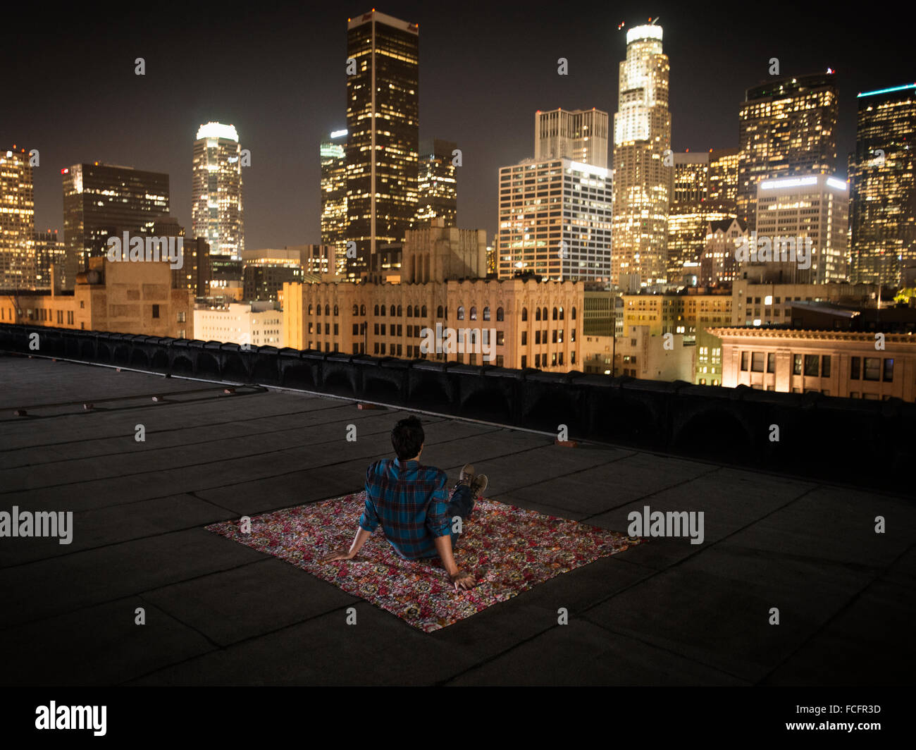 Ein Mann sitzt auf einem Teppich auf eine Dachterrasse mit Blick auf eine Stadt, die nachts beleuchtet. Stockfoto