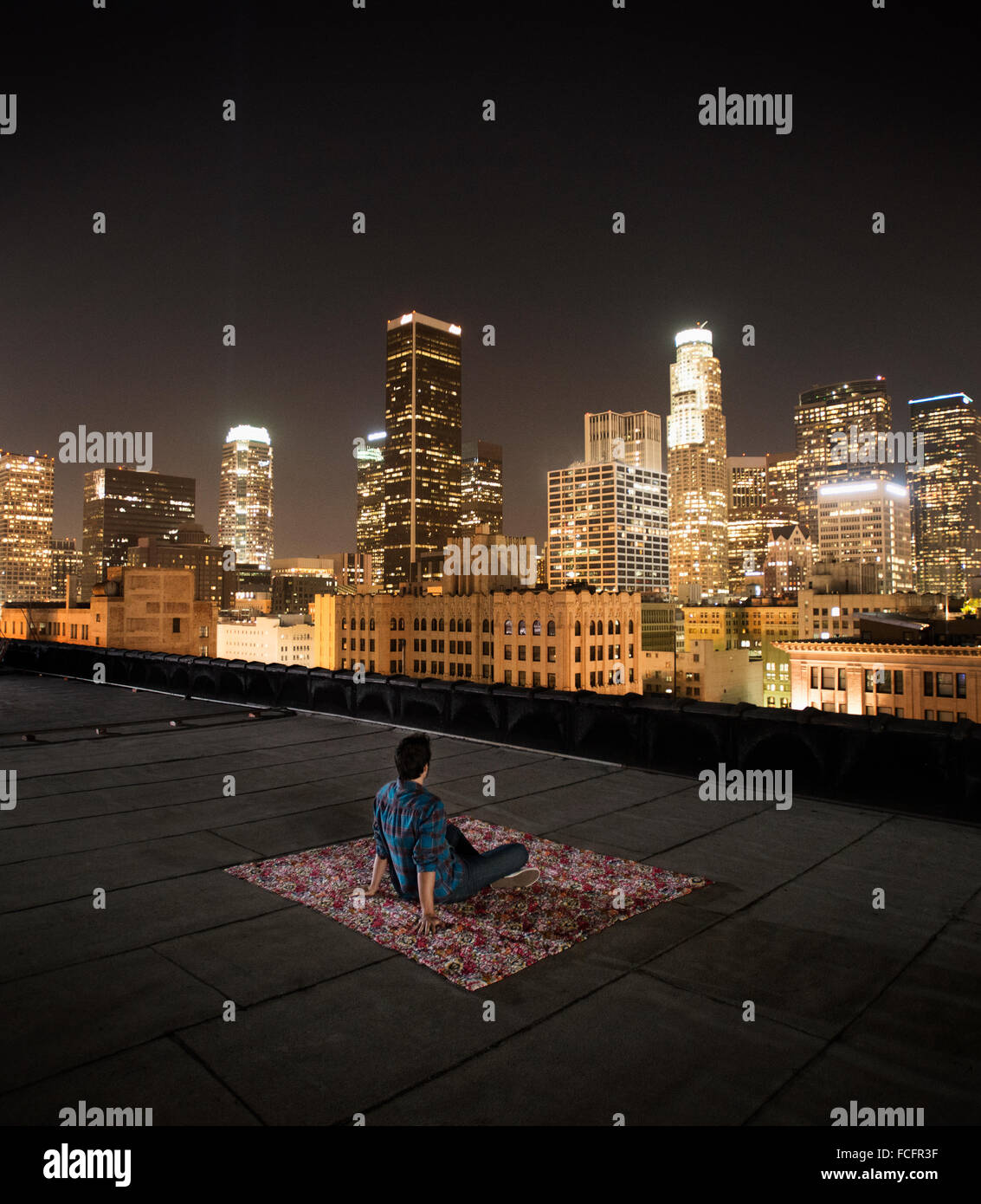 Ein Mann sitzt auf einem Teppich auf eine Dachterrasse mit Blick auf eine Stadt, die nachts beleuchtet. Stockfoto