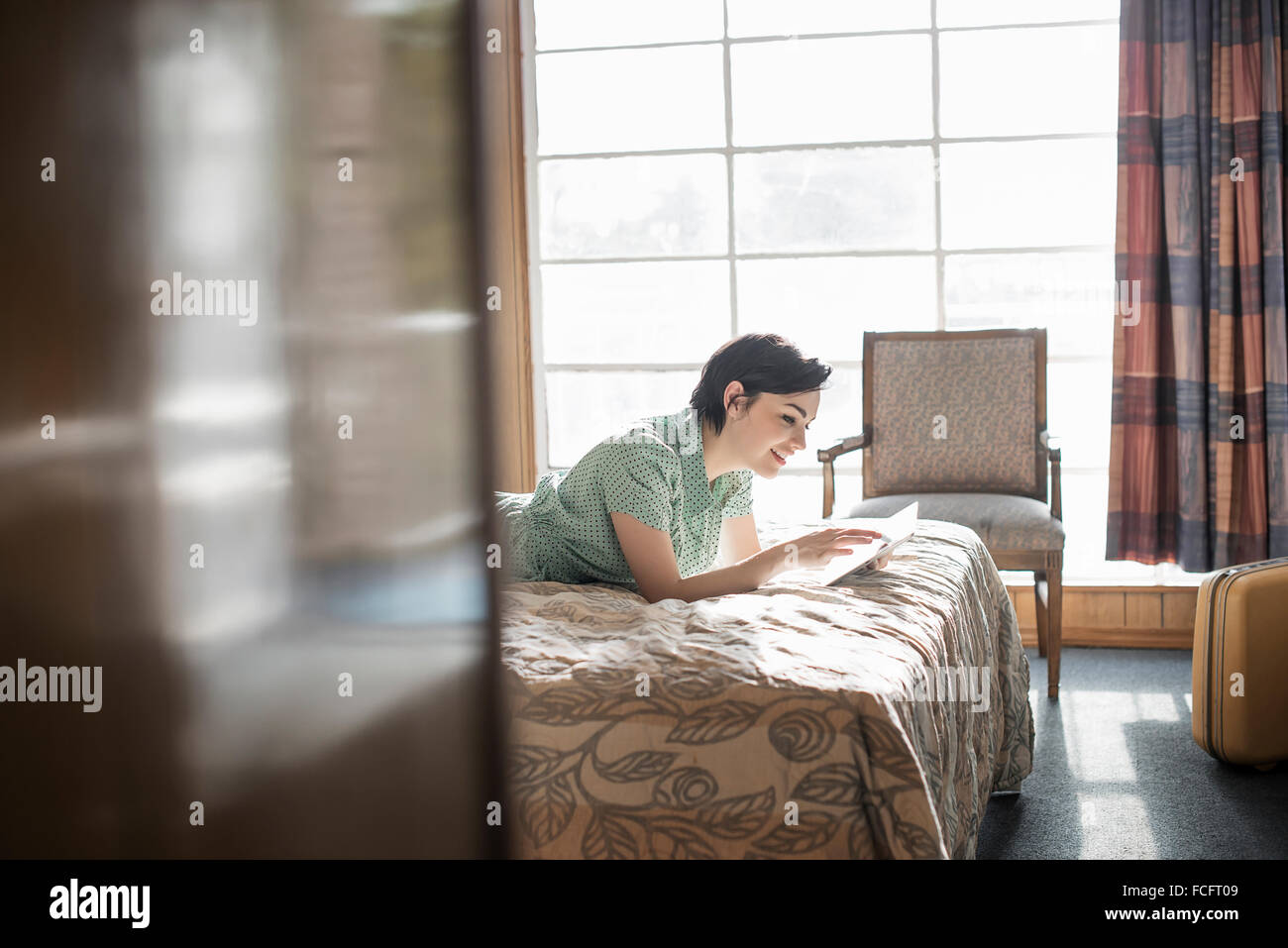 Eine junge Frau auf einem Bett in einem Motelzimmer mit einem digital-Tablette Touchscreen liegend überprüft Vorhänge, mobile Kommunikation Internet Stockfoto