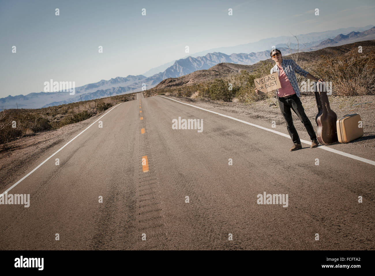 Ein Mann steht am Straßenrand, eine Anhalterin mit Gitarre und Fall hält ein Schild mit der Aufschrift Vegas oder pleite. Stockfoto