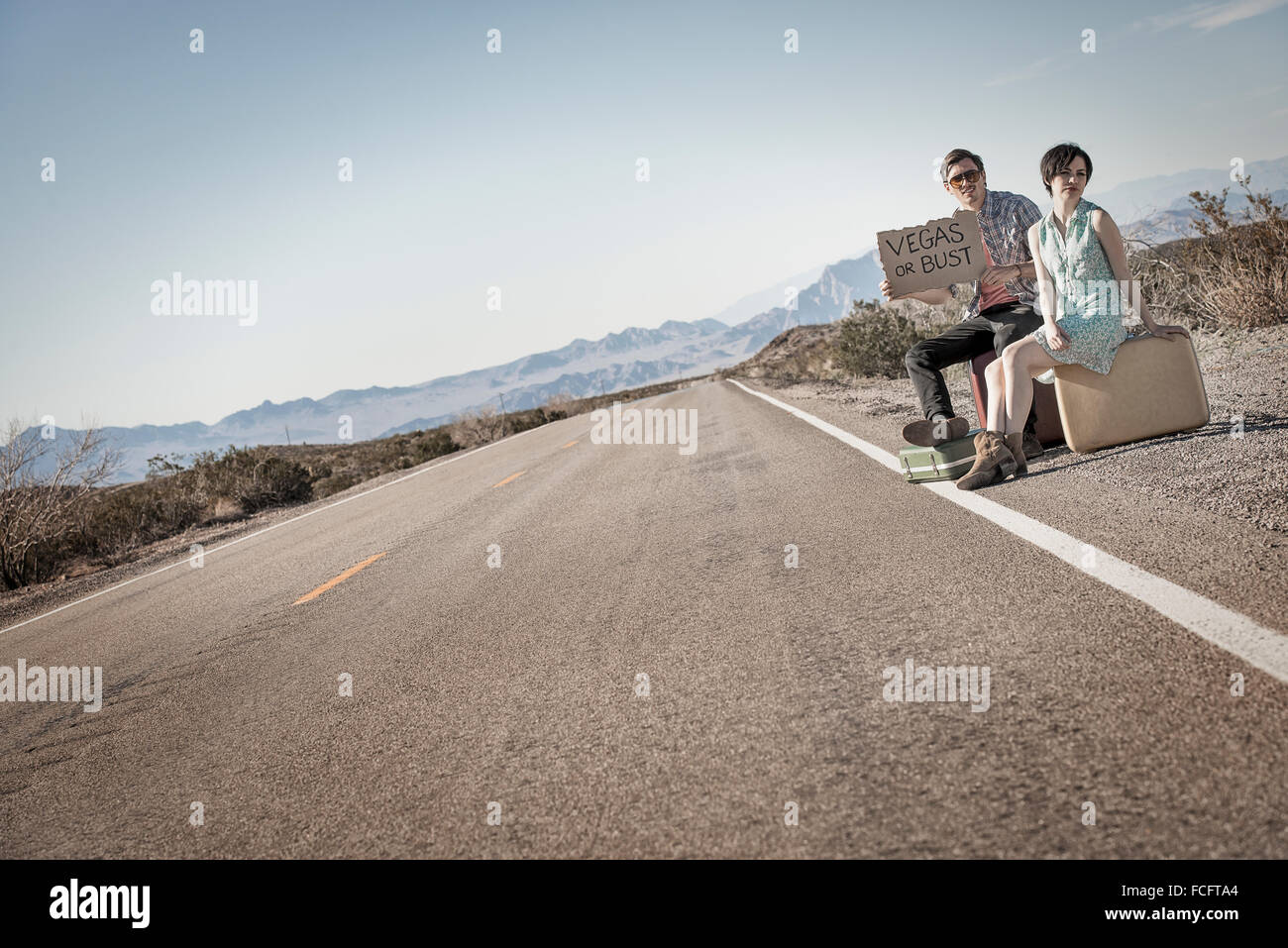 Ein junges Paar, Mann und Frau auf eine asphaltierte Straße in der Wüste Hitchiking, mit einem Schild mit der Aufschrift Vegas oder pleite. Stockfoto