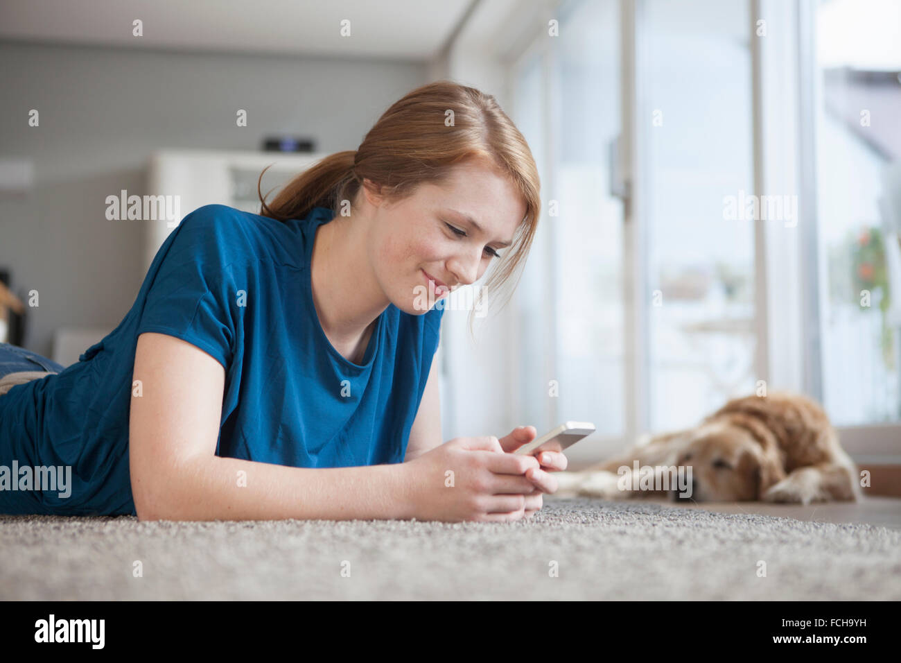 Lächelnde junge Frau liegt auf dem Boden im Haus auf der Suche auf smartphone Stockfoto