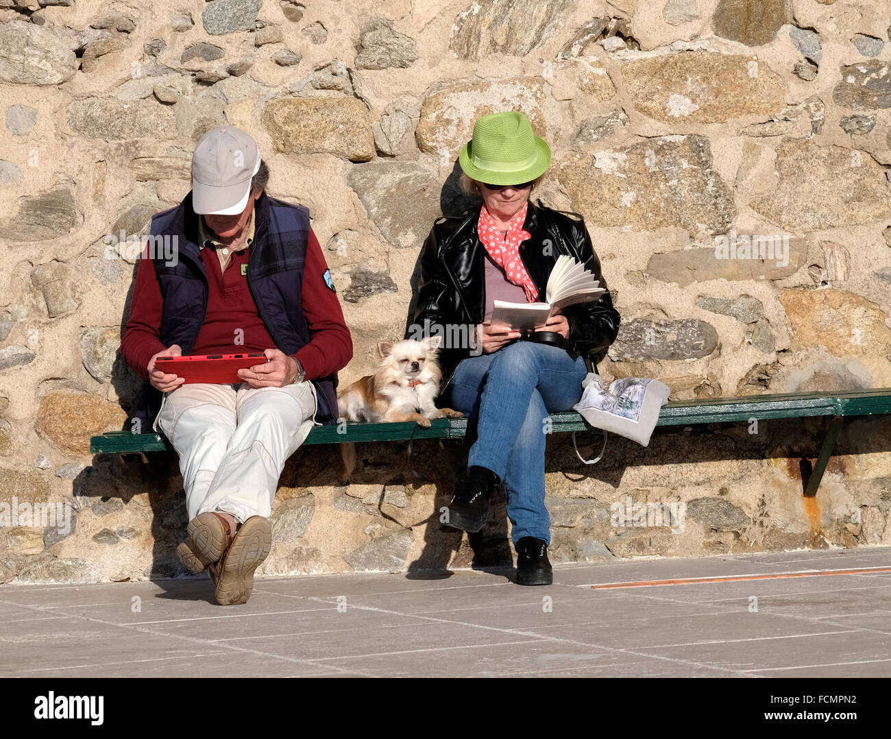 Applying paar im Urlaub in St. Tropez.  Beide sitzen, lesen, eines von einem Tablet, eine aus einem Taschenbuch.  Kleiner Hund zwischen. Stockfoto