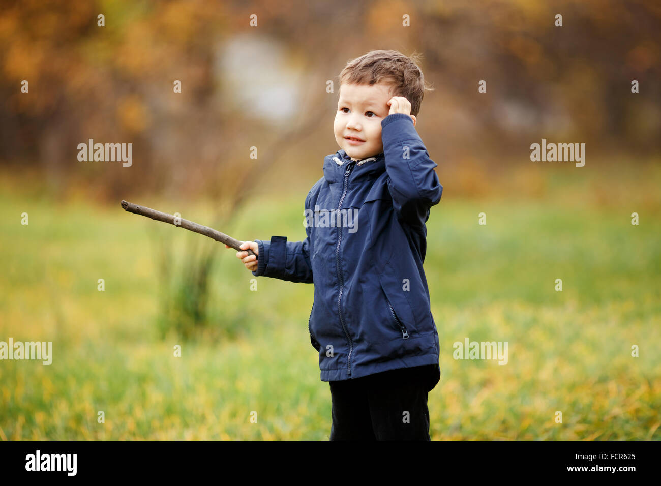 Netter Junge mit Holzstab in der Hand spielen im Herbst Park im Freien. Verwirrt, denken, was zu tun ist, auf der Suche zur Seite. Herbst, gelbe Blätter. Kinder tragen blaue Jacke. Unbeschwerte Kindheit. Stockfoto