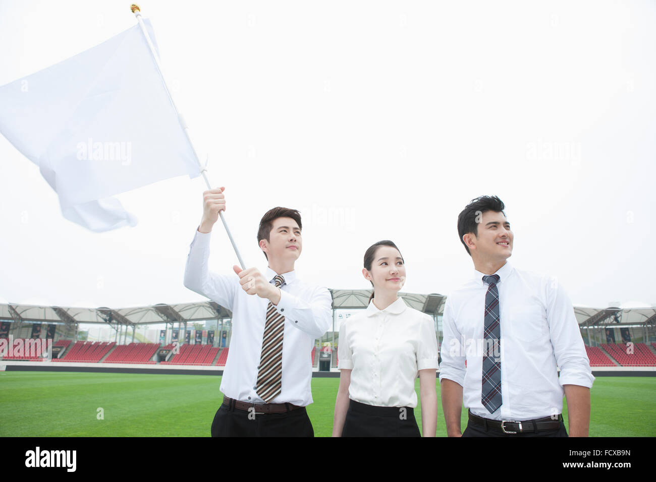 Drei junge Erwachsene in Business-Kleidung am Sportplatz hält eine weiße Fahne und starrte nach oben Stockfoto