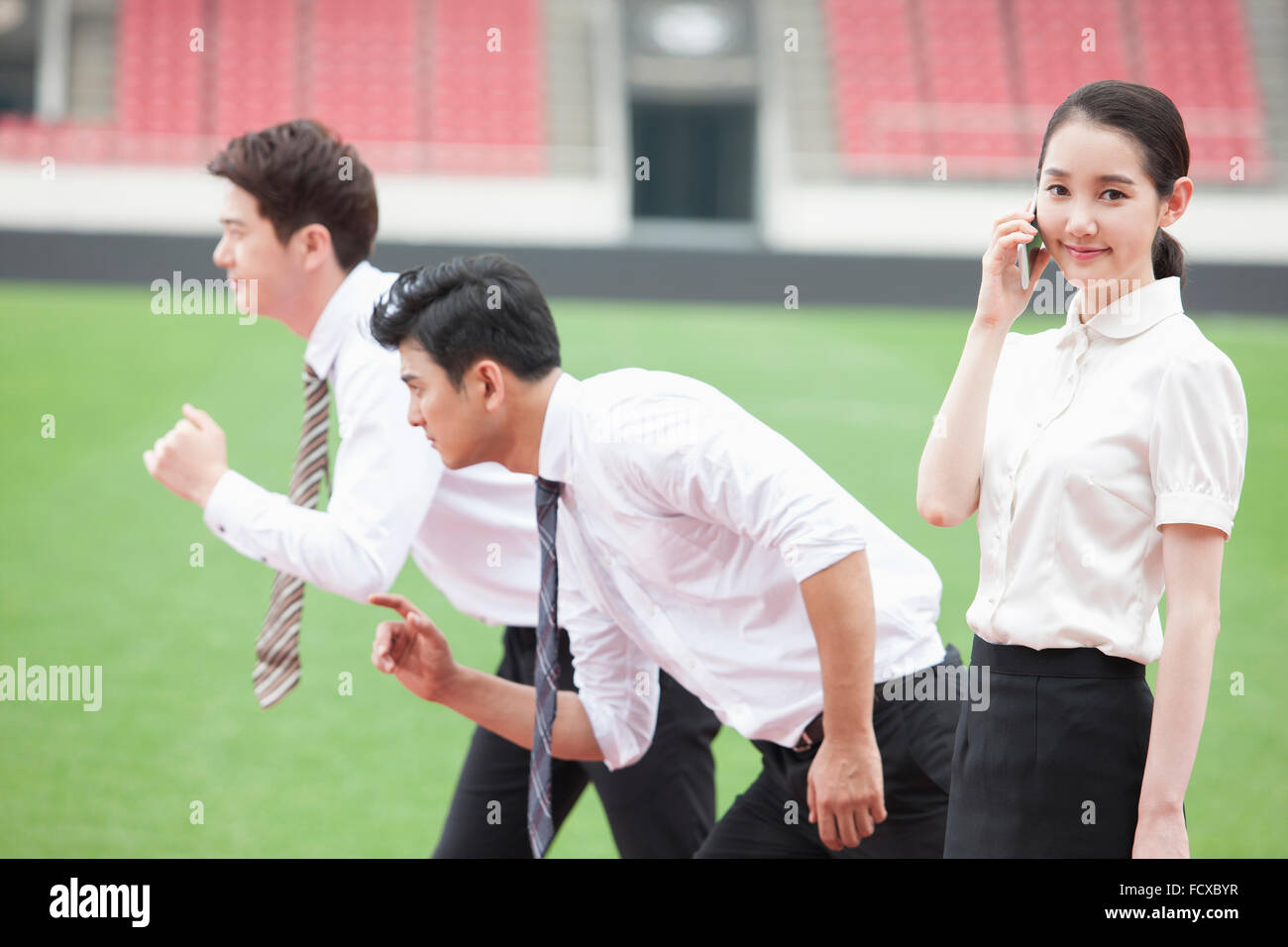 Zwei Männer in Business-Kleidung aufgereiht mit einer Pose für den Betrieb mit dem Hintergrund von Sportplatz und eine Frau auf ihrem Handy Stockfoto