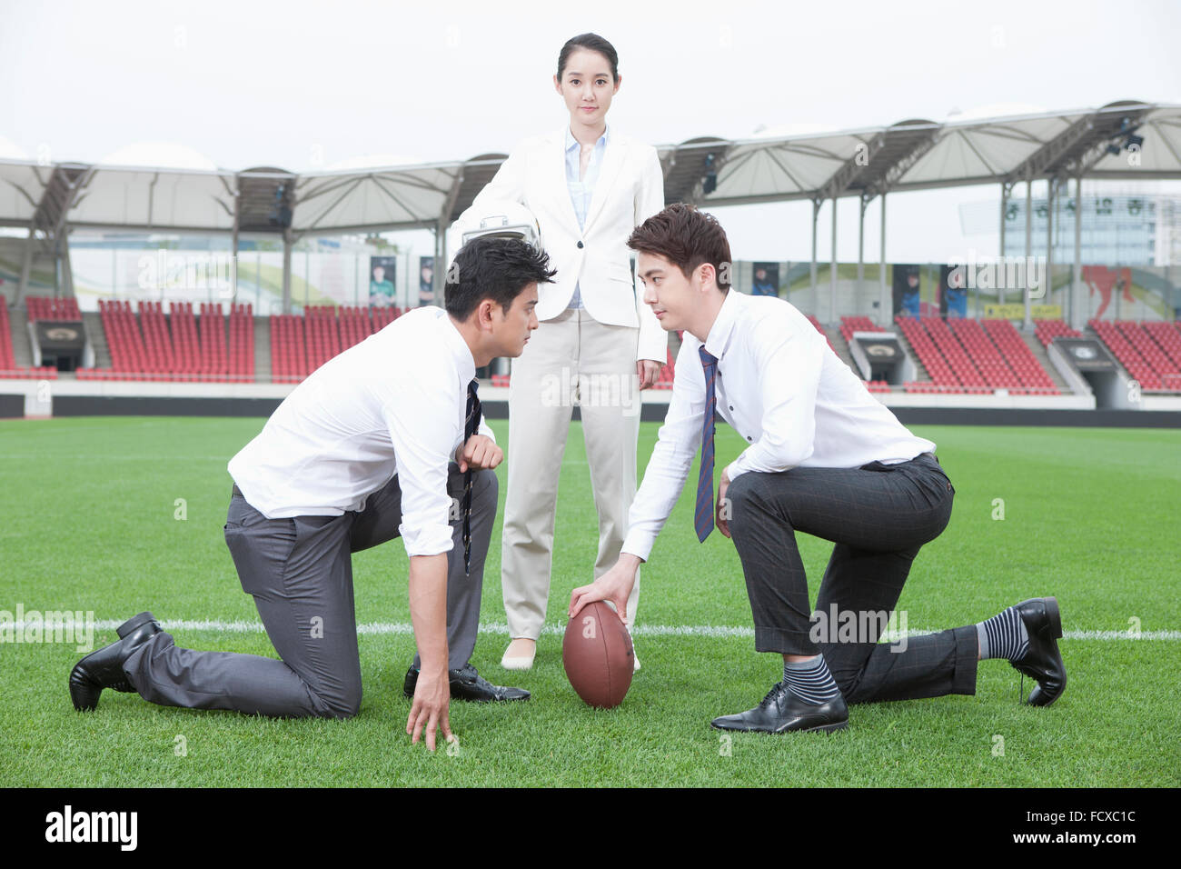 Zwei Geschäftsleute kniete auf Wiese mit einem Rugby-Ball mit eine Geschäftsfrau, die dazwischen stehen Stockfoto