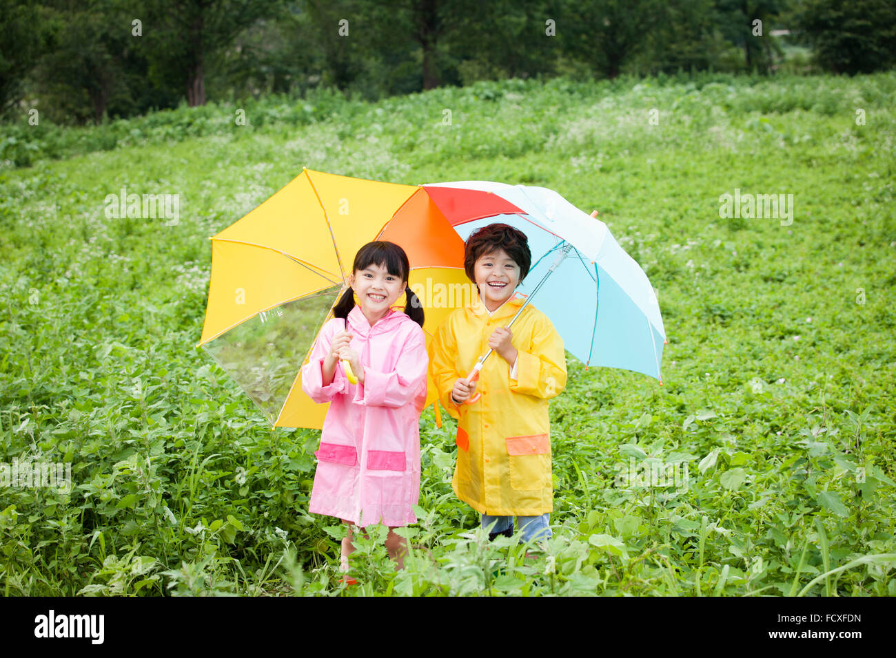 Junge und ein Mädchen im Regen Mantel unter einem Regenschirm am Feld und starrte nach vorne mit einem Lächeln zu jeder stehen Stockfoto