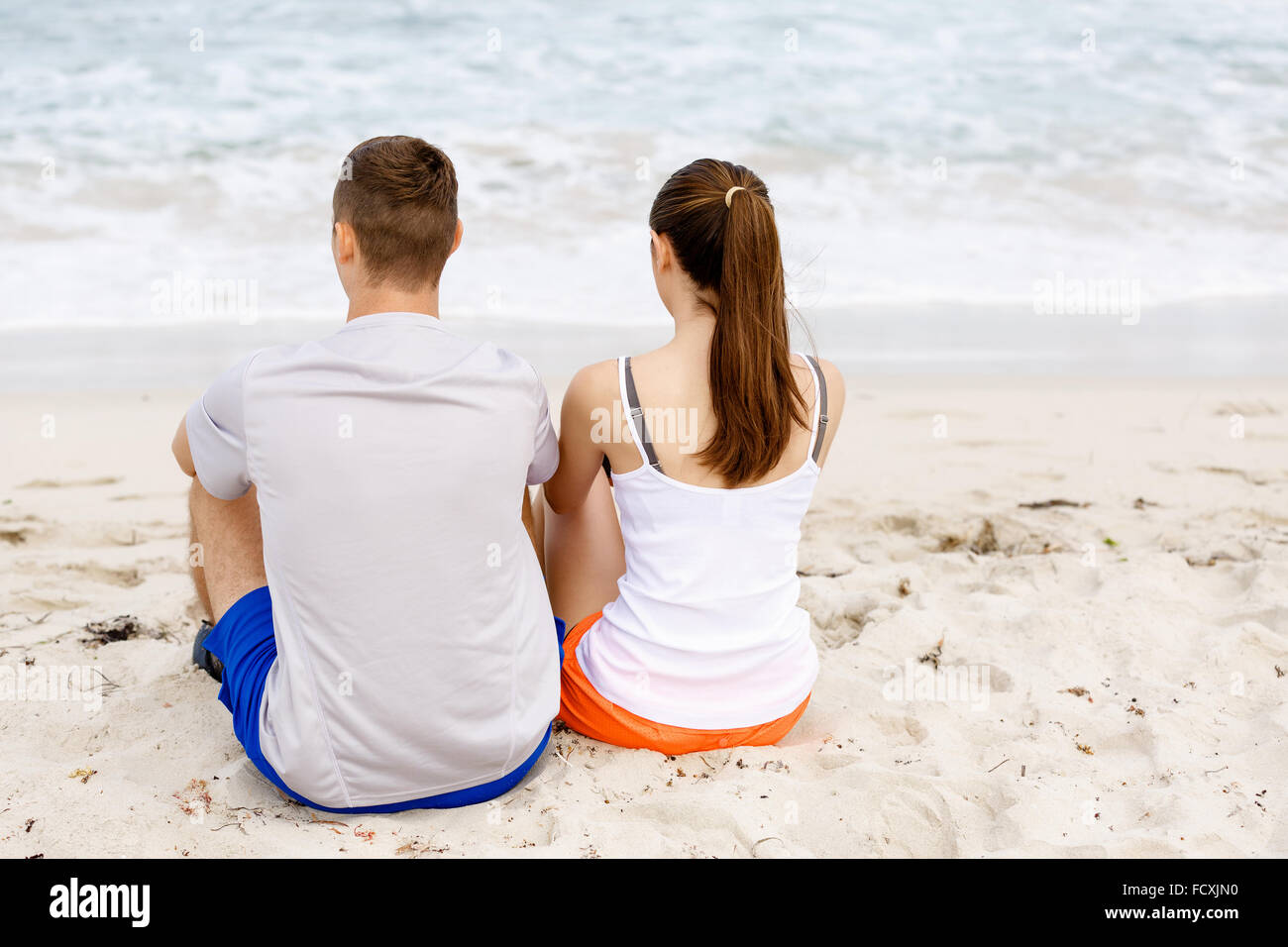 Junges Paar sahen einander sitzen zusammen am Strand in Sportbekleidung Stockfoto